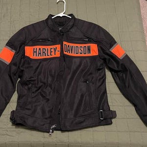 Könnte beinhalten: Eine schwarze Motorradjacke aus Mesh mit einem markanten horizontalen orangefarbenen Streifen auf der Brust, der den Schriftzug Harley-Davidson in schwarzen Buchstaben trägt. Die Jacke hat orangefarbene Ärmelbänder, Reißverschlusstaschen und verstellbare Seitengurte.