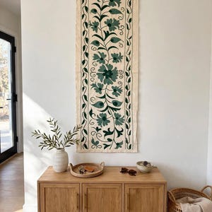 May include: A rectangular off-white fabric wall hanging featuring dark green embroidered floral and vine patterns. The textile is suspended from a wooden rod with a thin rope. The design includes symmetrical leaf motifs and central flower shapes.