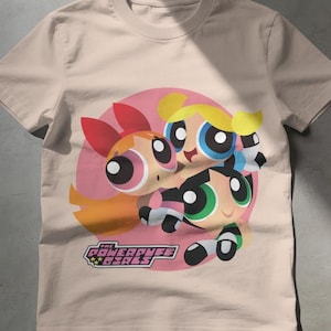 Puede incluir: Una camiseta beige de manga corta y cuello redondo con un gran gráfico circular de los tres personajes de Las Supernenas, Pétalo, Burbuja y Cactus, con el texto The Powerpuff Girls impreso en una fuente estilizada rosa y negra debajo.