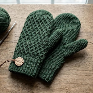 Op de afbeelding: Een paar donker bosgroene gehaakte wanten met een gestructureerd ruitpatroon op de achterkant en een geribbelde boord. Een rond houten label bevestigd met een bruin koord toont de tekst Alpine Texture Mittens Handmade Wool.