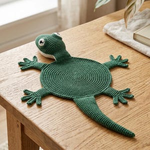 Op de afbeelding: Een donkergroene gehaakte mat in de vorm van een hagedis, met een cirkelvormig lichaam in een spiraalsteekpatroon, vier gespreide poten, een lange puntige staart en een kop met wit-zwarte ogen. Het item is gemaakt van dicht, gestructureerd garen.