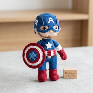 Könnte beinhalten: Eine gehäkelte Captain America Puppe mit einer blauen Maske mit weißem A, einem rot-weiß gestreiften Oberkörper mit weißem Stern, blauen Beinen und roten Stiefeln. Die Puppe hält einen runden rot-weiß-blauen Schild. Ein Holzblock trägt die Aufschrift Kashmala Stitches.