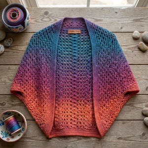May include: A crochet shrug featuring an openwork stitch pattern with a gradient color transition from orange at the bottom to purple in the center and blue at the top. A brown rectangular label with the text Rhythm Story is sewn at the neckline.
