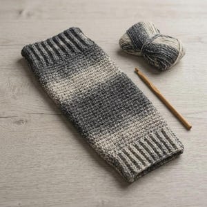 Puede incluir: Una pieza rectangular de ganchillo con un degradado de hilo gris carbón a crema claro con bordes acanalados en ambos extremos. Junto a la tela hay un ovillo de hilo jaspeado a juego y un ganchillo de madera para proyectos de manualidades.
