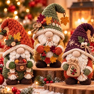 May include: Three handcrafted crochet gnomes featuring autumn-themed attire. The left gnome wears a rust-colored hat with acorns, the center gnome a green hat with leaves, and the right gnome a purple hat with a star, all with white beards.