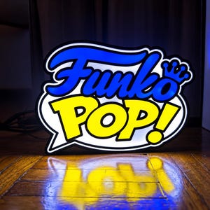 Può includere: Un'insegna luminosa Funko Pop! che mostra il nome del marchio in lettere blu in grassetto con una piccola icona a forma di corona blu sopra la lettera O, su uno sfondo a fumetto giallo brillante con un bordo nero spesso.