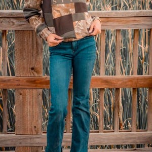 May include: A long-sleeve hooded sweatshirt featuring a patchwork design of brown solid fabric and camouflage print fabric with the text SITKA printed on the chest, paired with blue denim jeans and brown leather boots with a decorative stitched pattern.