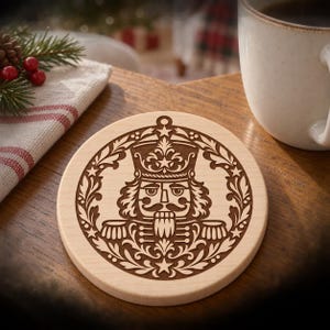 Puede incluir: Un posavasos redondo de madera clara con un diseño grabado a láser de un cascanueces centrado dentro de un marco circular de hojas ornamentadas y pequeñas estrellas. La figura del cascanueces lleva un sombrero alto y un uniforme detallado.