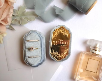 May include: A light blue velvet ring box containing three gold rings, with a gold mirror lid engraved with "THE MILLERS EST. 2016". The set is accompanied by a glass perfume bottle, a spool of light blue silk ribbon, and a bouquet of pale pink flowers.