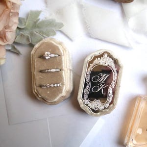 May include: A beige velvet ring box with three silver rings and a matching lid featuring an ornate silver frame with the engraved letters MK. The box is positioned next to a clear glass perfume bottle with a gold cap and a bouquet of light flowers.