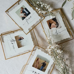 May include: Four gold-framed table numbers with photos and torn-edge paper. Each frame has a different photo of a couple and a number written in cursive. The names "Natalie & Edward" and the date "10.12.24" are also written on each card.