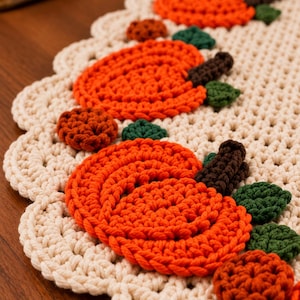 May include: A cream-coloured crocheted table runner featuring a scalloped edge and a repeating pattern of orange pumpkins with dark brown stems and small dark green leaves. The pumpkins are textured and stitched directly onto the base fabric.