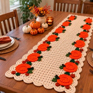 Puede incluir: Un camino de mesa rectangular de ganchillo en color crema con un borde festoneado y un patrón repetitivo de calabazas de color naranja brillante con hojas verde oscuro y pequeñas bayas rojo oscuro a lo largo de la pieza.