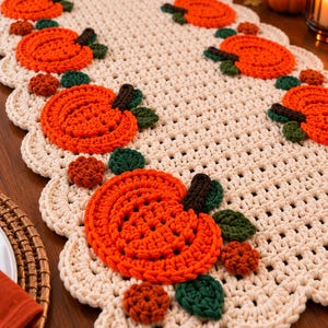 May include: A cream-coloured crocheted table runner featuring a scalloped edge and a repeating pattern of bright orange pumpkins with dark brown stems and dark green leaves, accented by small, round, textured orange and brown floral motifs.