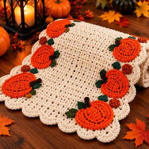 May include: A cream-coloured crocheted table runner featuring a scalloped edge and decorative appliqu&eacute;s of bright orange pumpkins with dark green stems and leaves. The runner is rolled at one end and displays a repeating pattern of pumpkins.