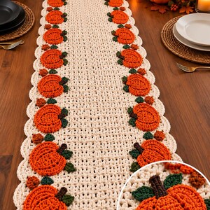 May include: A cream-coloured crocheted table runner featuring a repeating pattern of orange pumpkins with dark green leaves and brown stems along both scalloped edges. The runner has a textured, open-weave design with a circular inset detail.