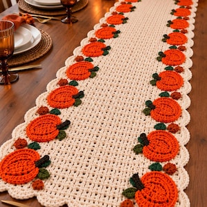 May include: A cream-coloured crocheted table runner featuring a repeating pattern of orange pumpkins with dark green leaves and small brown accents along the scalloped edges. The runner has an open-weave grid design and a decorative scalloped border.