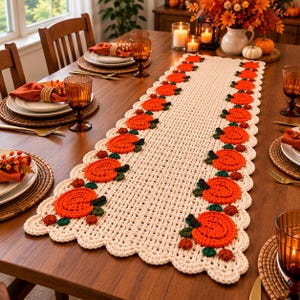 May include: A cream-coloured crocheted table runner featuring a repeating pattern of orange pumpkins with green stems and leaves along both sides. The runner has a scalloped edge and a grid-like centre design, laid flat on a wooden dining table.