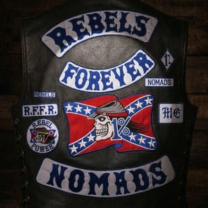 May include: A black leather vest featuring embroidered patches. The back displays the text REBELS FOREVER and NOMADS in blue and white. A central patch shows a skull wearing a hat over a red, white, and blue flag with the text 1% and REBEL POWER.