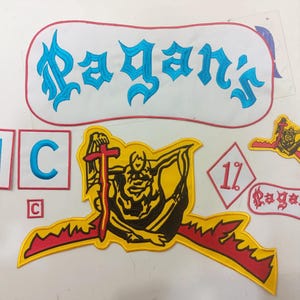 Puede incluir: Una colección de parches bordados que incluye un parche grande de color blanco con el texto Pagan's en azul, dos parches cuadrados con las letras MC en azul, un parche grande de figura amarilla y negra con una espada roja y un parche de diamante 1%.