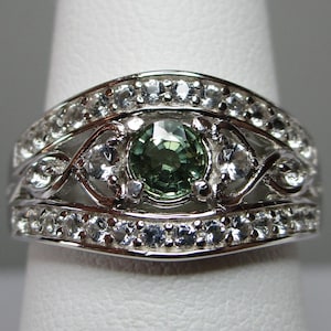 May include: A silver ring featuring a central green gemstone, encircled by smaller clear stones. The ring showcases an intricate, decorative pattern, with a band of clear stones on each side of the central design. The ring is presented on a white finger form.