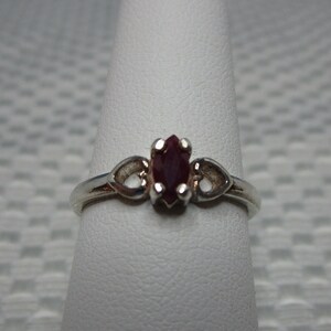 May include: A silver ring featuring a maroon oval-cut gemstone. The ring has a delicate design with decorative swirls on either side of the gemstone. The ring is displayed on a white finger form.