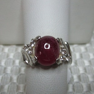 May include: A silver ring featuring a large, oval, deep red gemstone. The ring's band has an ornate design with small, clear accent stones. The ring is displayed on a white stand against a textured background.
