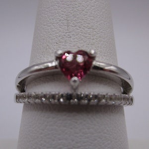 May include: A silver ring with a heart-shaped, red gemstone as the centerpiece. Below it, a second ring with small, clear stones encircles the band. The rings are displayed on a white, conical stand, showcasing the jewelry's design.