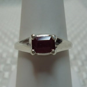 May include: A silver ring with a rectangular, deep red gemstone. The ring's band is simple, with a slight taper towards the gemstone. The gemstone is set in a four-prong setting, and the ring is displayed on a white stand.