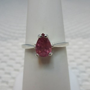 Puede incluir: Anillo de plata con una gema rosa en forma de pera. La gema destaca por su engaste sencillo y elegante. El anillo se exhibe sobre un soporte blanco, resaltando su diseño y color.