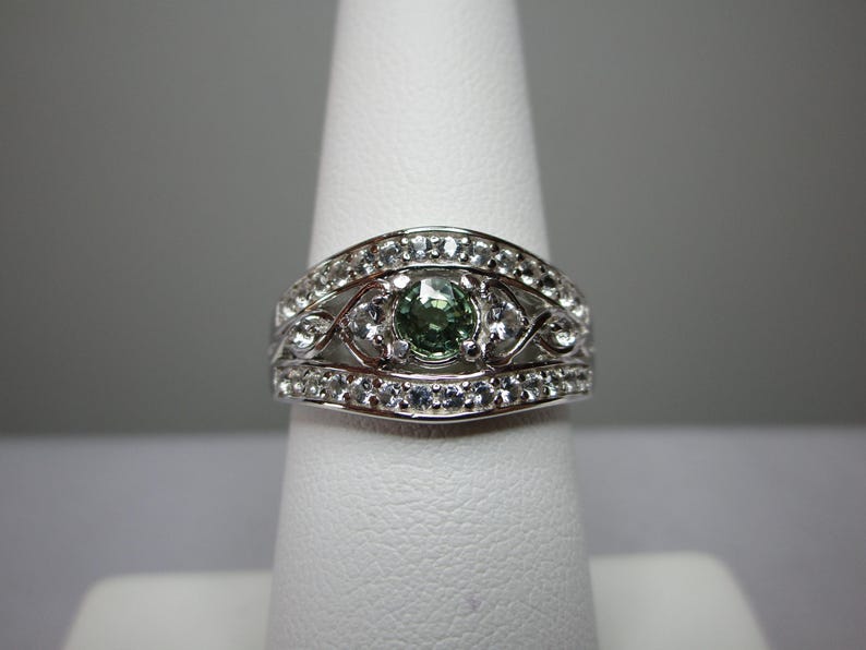 May include: A silver ring featuring a central green gemstone, encircled by smaller clear stones. The ring showcases an intricate, decorative pattern, with a band of clear stones on each side of the central design. The ring is presented on a white finger form.