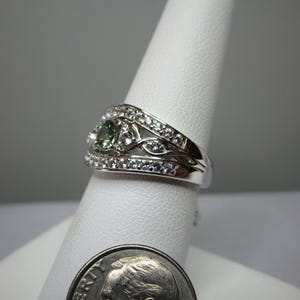 May include: A silver ring featuring a central green gemstone and many small, clear stones. The ring is displayed on a white stand, with a dime in the foreground for scale. The design incorporates floral and leaf-like patterns.