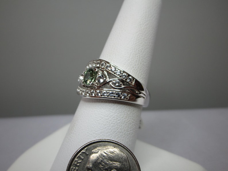 May include: A silver ring featuring a central green gemstone and many small, clear stones. The ring is displayed on a white stand, with a dime in the foreground for scale. The design incorporates floral and leaf-like patterns.