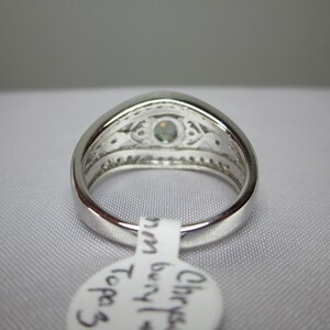 May include: A silver ring featuring a light-green gemstone at its centre. The ring has an intricate design around the gem and along the band. A white tag with text is attached.