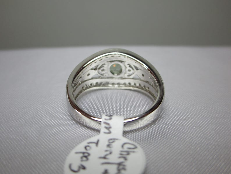 May include: A silver ring featuring a light-green gemstone at its centre. The ring has an intricate design around the gem and along the band. A white tag with text is attached.