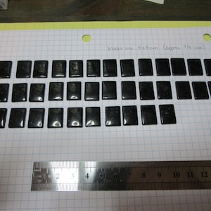 May include: A collection of rectangular, glossy black blocks, each approximately 2 inches long and wide, arranged on graph paper. The text indicates the blocks are 5x5cm (approx. 3/8 inch). A metal ruler is positioned below the blocks.