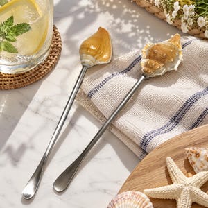 May include: Two long-handled metal stirring spoons featuring decorative natural seashell finials. One spoon is topped with a smooth, tan, cone-shaped shell, while the other features a textured, brown and cream-colored spiral seashell.
