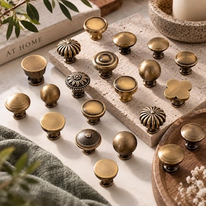 May include: A collection of antique brass cabinet knobs in various shapes and textures, including smooth round, fluted, and floral designs. These metal hardware pieces feature a weathered, aged finish and are arranged on a stone surface.