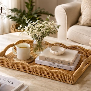 Puede incluir: Una bandeja rectangular de ratán trenzado con bordes festoneados y asas laterales. Dentro, una taza de cerámica blanca descansa sobre un pequeño posavasos, junto a un jarrón de cristal transparente con flores blancas, un cuenco blanco pequeño y dos libros apilados titulados Live Beautifully y This Is Home.