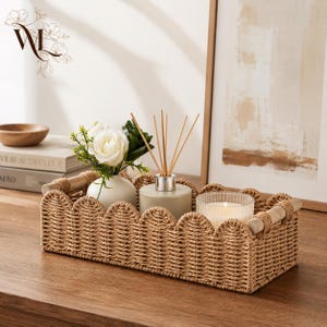 May include: A rectangular woven basket with a scalloped top edge and light wood handles. Inside the basket sits a small white vase with white artificial flowers, a beige reed diffuser with wooden sticks, and a ribbed glass candle.