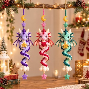 May include: Three hanging crochet dragon ornaments in purple, pink, and teal. Each dragon features a coiled tail, wings, and holds a small object. Above each dragon is a yellow crescent moon and a star, with a white cloud and star at the bottom.