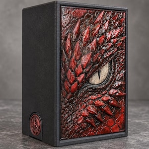 Può includere: Una scatola rettangolare nera dotata di un rilievo tridimensionale di un occhio di drago rosso sul pannello frontale. La texture della pelle di drago è dettagliata con squame appuntite e in rilievo che circondano un occhio centrale di colore chiaro.