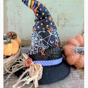 May include: A black Halloween witch hat with a wide brim, decorated with a white spiderweb design, orange cross stitches, and a purple crescent moon. The hat features a blue polka dot ribbon and a large wooden button with raffia. A festive decoration.