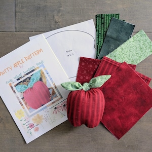 May include: A handmade red apple with green leaves sits next to fabric squares and a pattern. The pattern is for a "Knotty Apple" and includes the text "Look What Summer Made". Various fabric pieces in red and green are also included.