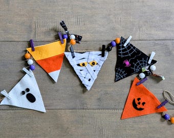 May include: A Halloween-themed banner featuring felt triangle flags. Designs include a candy corn, mummy, spiderweb, ghost, and jack-o'-lantern. Each flag is attached to a string with small clothespins and decorative beads. The banner is set against a wooden background.