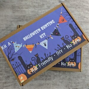 May include: Two cardboard boxes with a purple background featuring a Halloween bunting kit. The boxes have the words "Halloween Bunting Kit" printed on them, along with illustrations of pumpkins, cats, and bats. The kit is kid-friendly and no-sew.