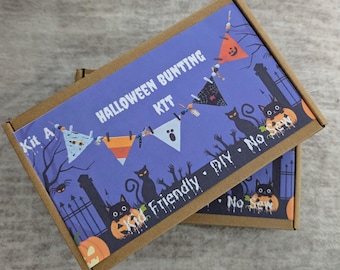 May include: Two cardboard boxes with a purple background featuring a Halloween bunting kit. The boxes have the words "Halloween Bunting Kit" printed on them, along with illustrations of pumpkins, cats, and bats. The kit is kid-friendly and no-sew.