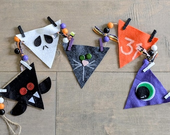 May include: A Halloween-themed banner featuring felt triangle pennants with various designs. The pennants include a ghost, a cat face, the number 31, a bat, and an eye. Each pennant is decorated with buttons, beads, and clothespins, creating a festive decoration.
