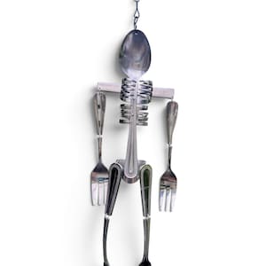 May include: A hanging sculpture constructed from stainless steel cutlery, featuring a spoon as the head, a horizontal metal bar for shoulders, and a ribcage made of bent fork tines. The arms and legs are formed from various fork and spoon handles.
