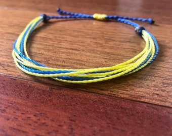 May include: A braided bracelet in yellow and blue, lying on a wooden surface. The bracelet's design is made of multiple threads. The colours are bright and the bracelet is adjustable.